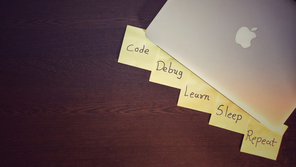 Laptop mit Notizzetteln „Code, Debug, Learn, Sleep, Repeat“ als Symbol für Bootcamp-Schüler, Quereinsteiger und Junior-Entwickler auf dem IT-Arbeitsmarkt
